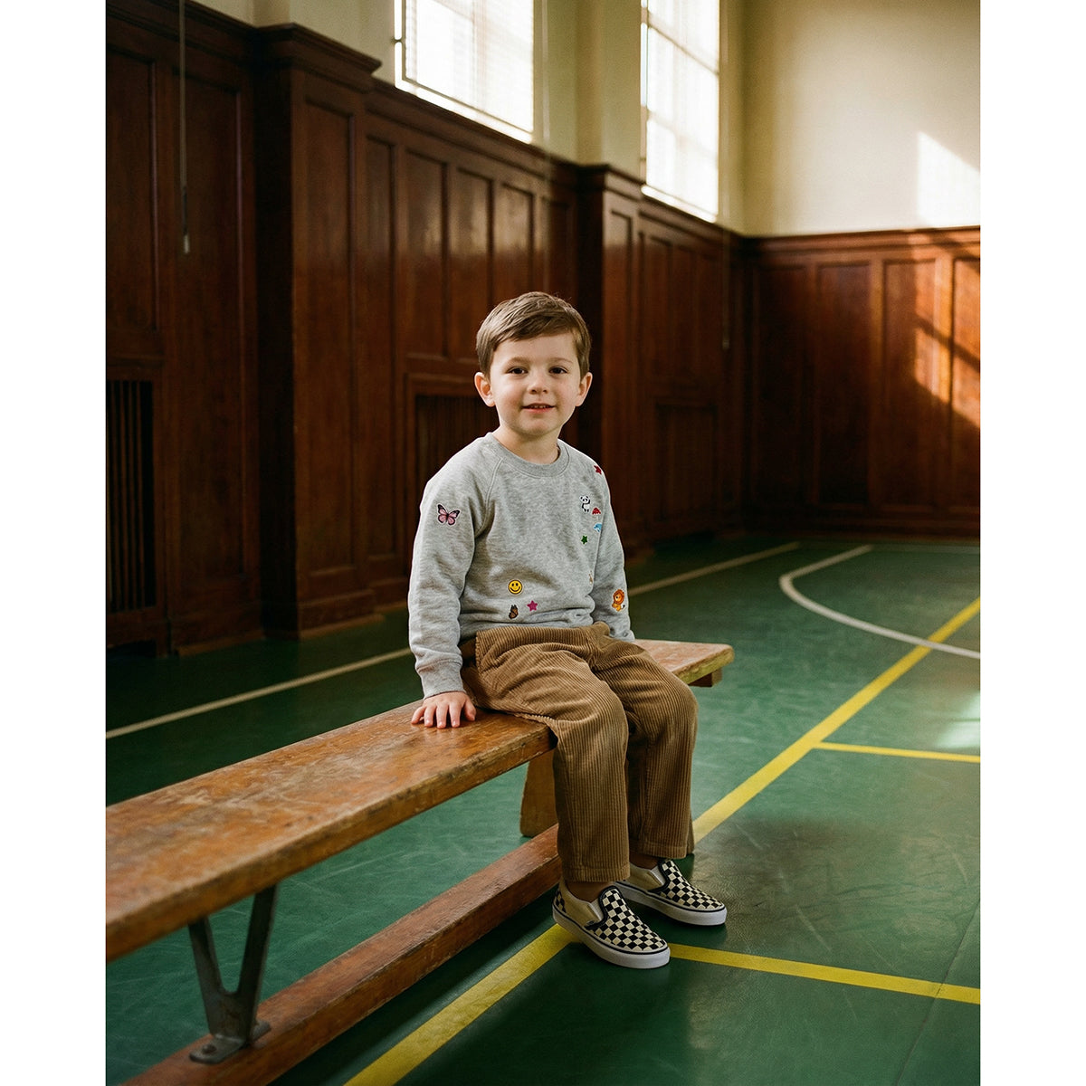 Child sitting on a wooden bench in a room with wooden paneling and green floor wearing a heather grey sweatshirt with printed stickers from BIKIND