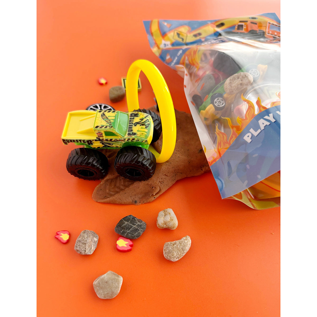 Earth Grown KidDoughs Toy truck on a textured brown KidDough with a yellow drive-through ring of fire toy on an orange surface, surrounded by small stones, fire fimos and the left side of the PlayKit packaging.
