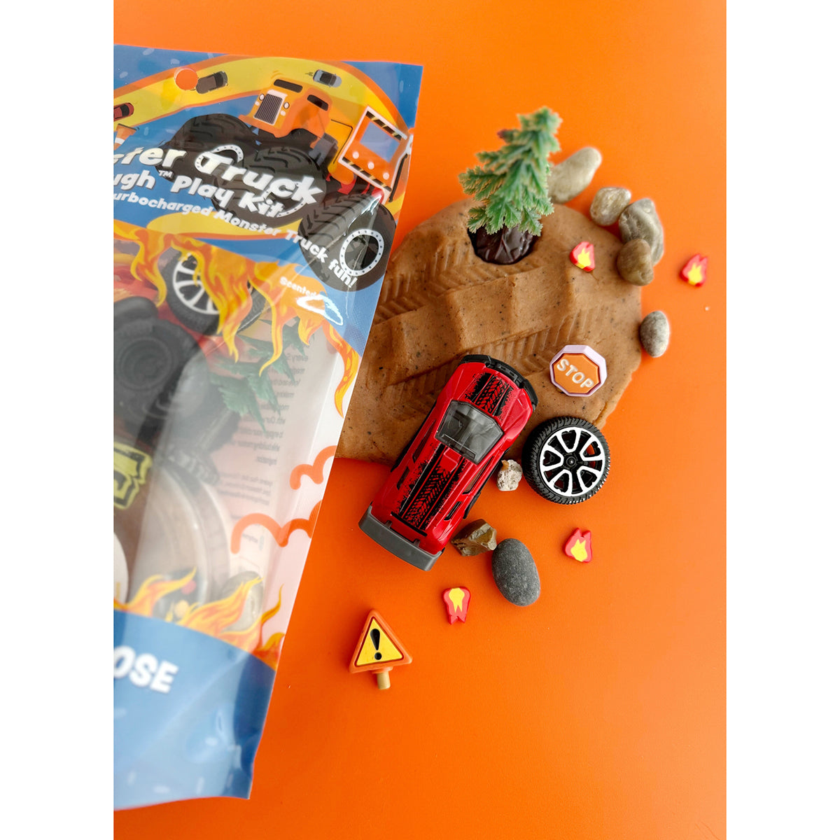 A miniature landscape with brown textured KidDough, toy truck, fire fimos, traffic signs, tire and natural rocks on an orange background with the right side of the Earth Grown KidDoughs Monster Truck PlayKit packaging next to it.