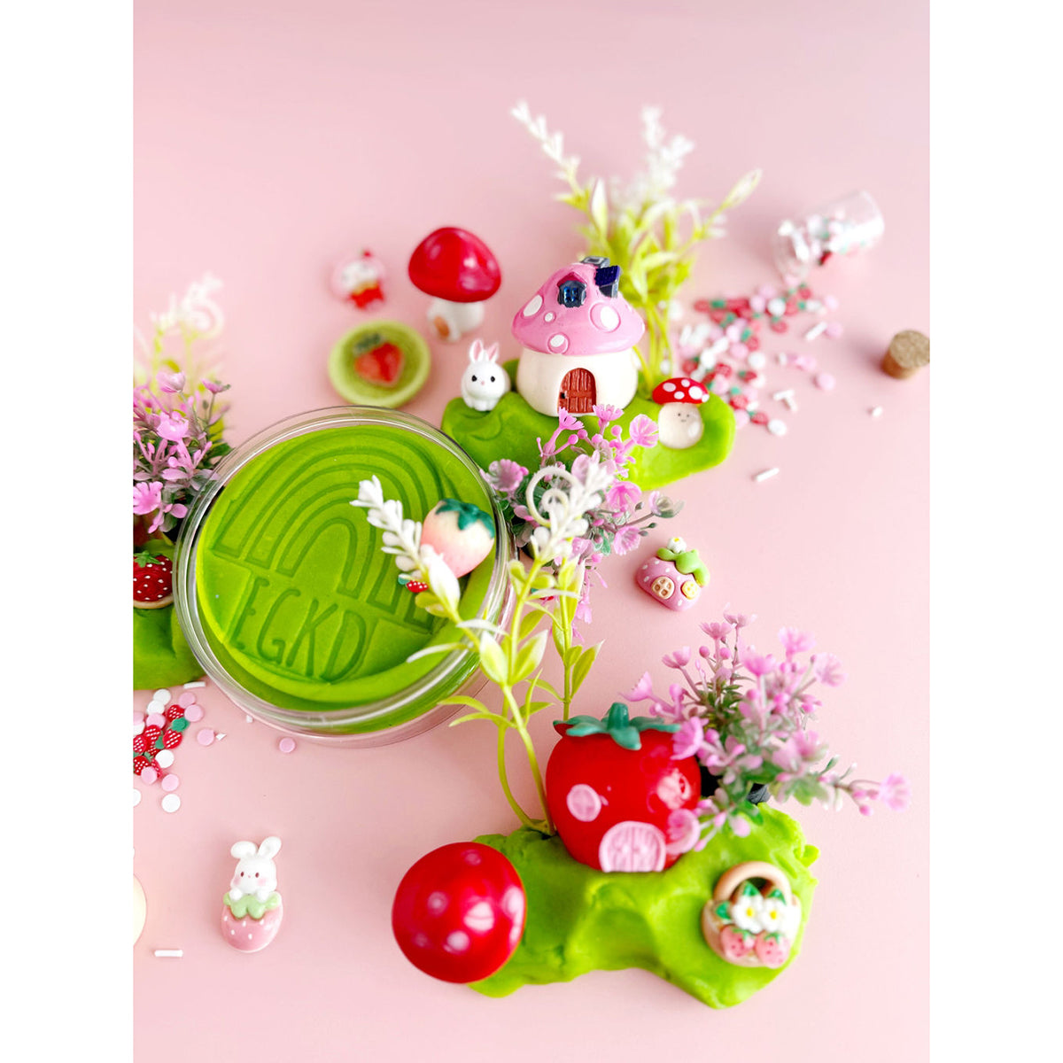 Earth Grown KidDoughs Bunny Berry Village KidDough Sensory Play Kit on a pink background. Close-up of of a jar with green KidDough and toy play pieces scattered around it.