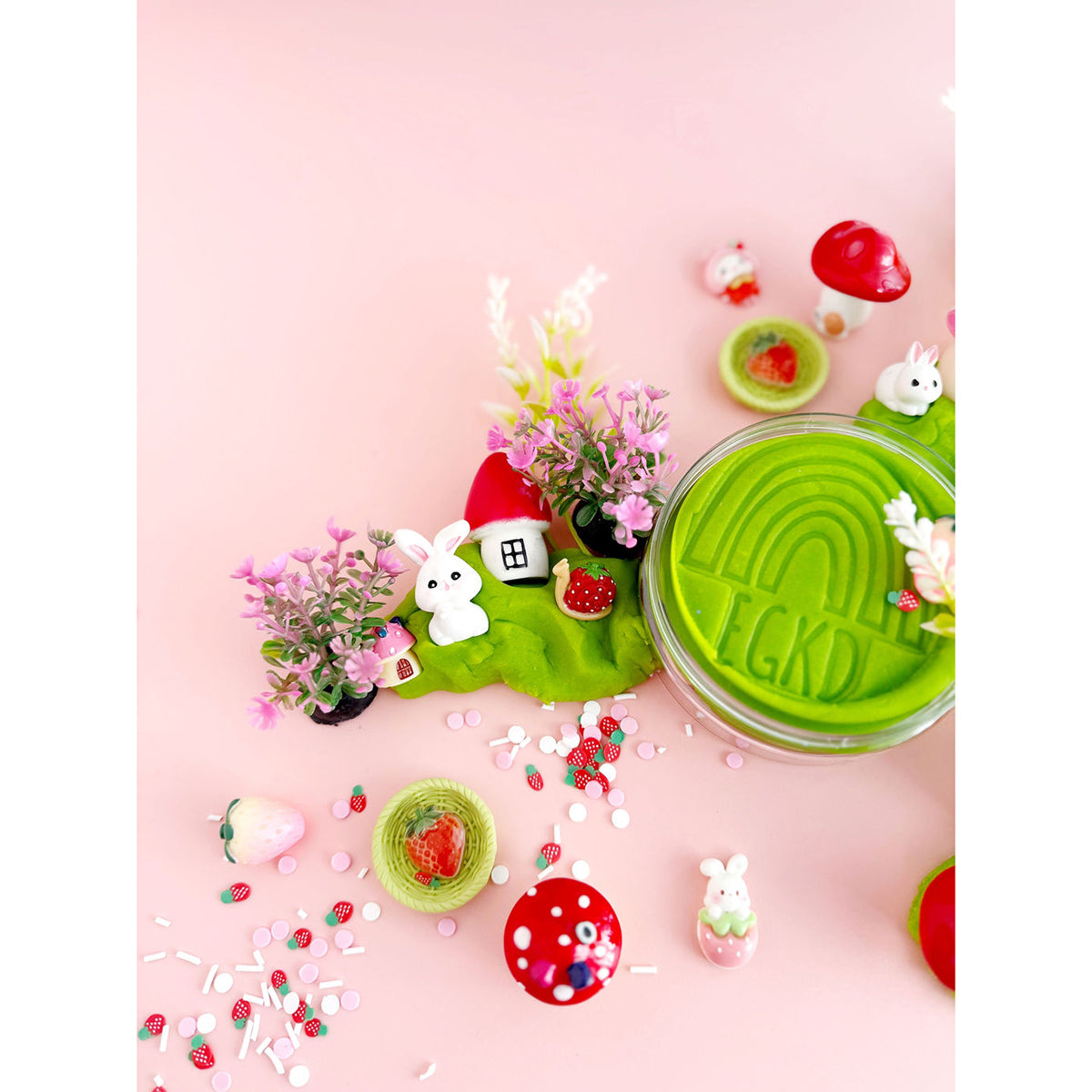Earth Grown KidDoughs Bunny Berry Village KidDough Sensory Play Kit on a pink background. Close-up of of a jar with green KidDough and toy play pieces scattered around it.