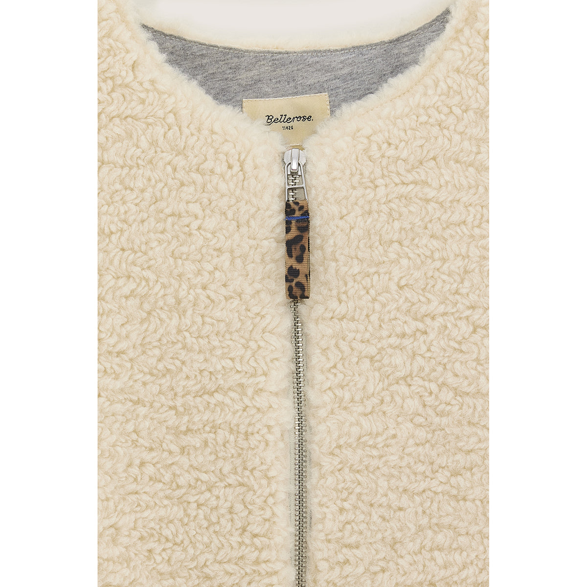Close-up product picture of Jams Bodywarmer front side from Bellerose. Bodywarmer is made from off-white teddy fabric, featuring a light grey fleece lining and has a front zipper closure with a leopard printed pull tab.